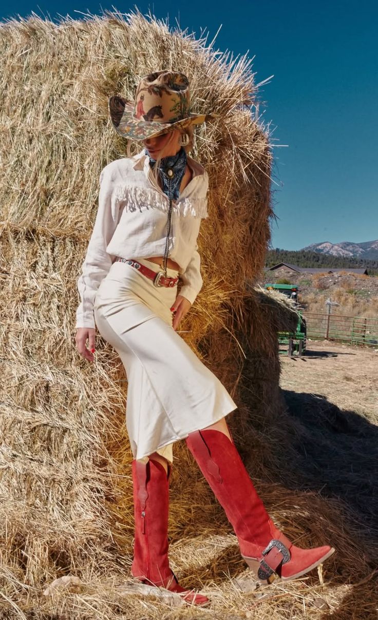 Woman in a cowboy hat and red boots leaning against hay bales outdoors