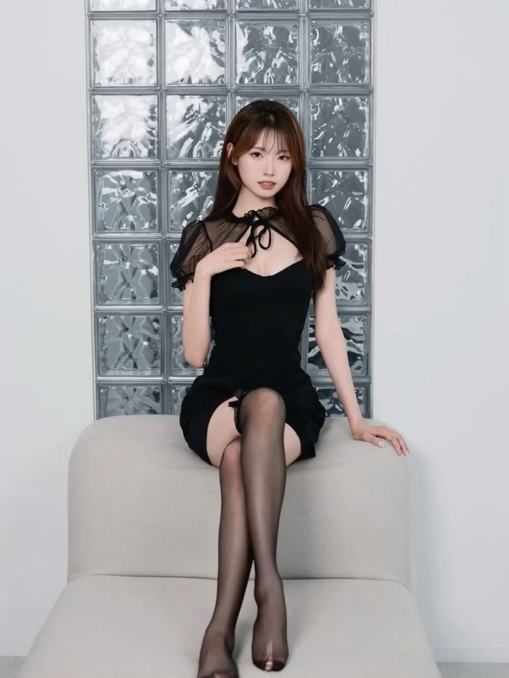 Woman in a black dress and stockings sitting in front of a glass block wall