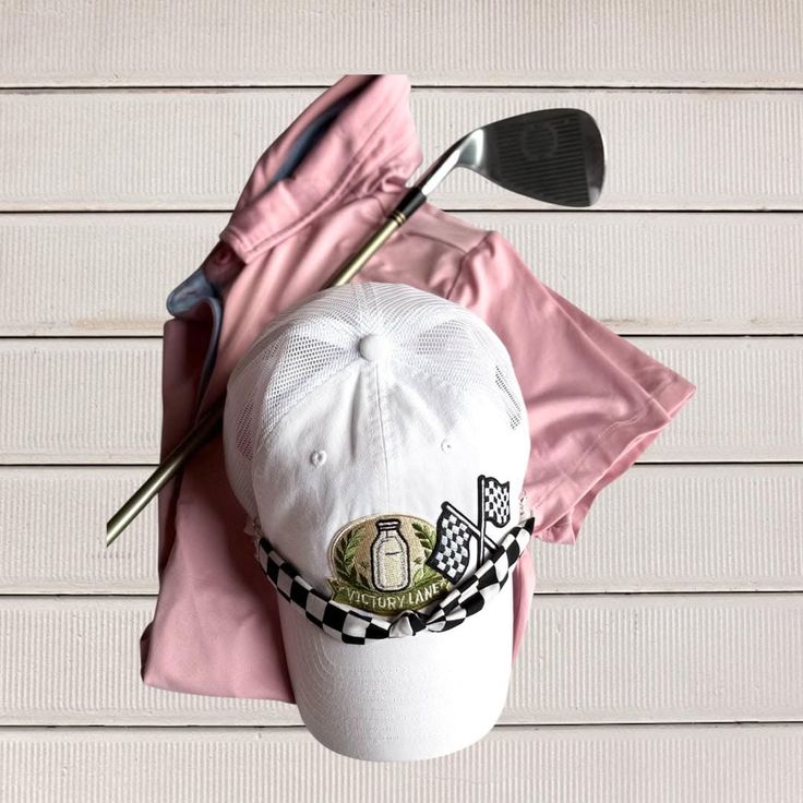 Golf club, white hat with checkered ribbon, and pink shirt on a white wooden surface