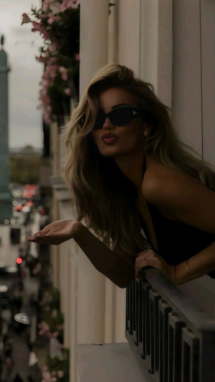 Blonde woman in sunglasses blowing a kiss from a balcony overlooking a city