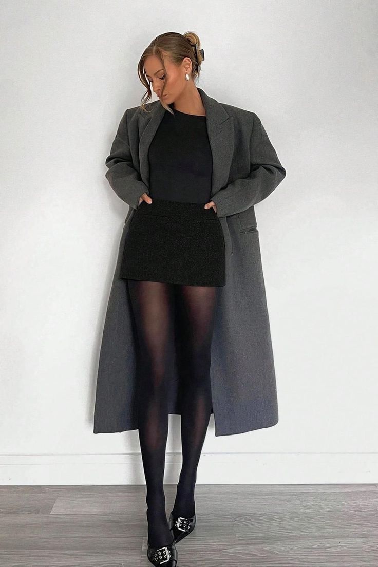 Woman in a long gray coat, black skirt, and black tights posing indoors