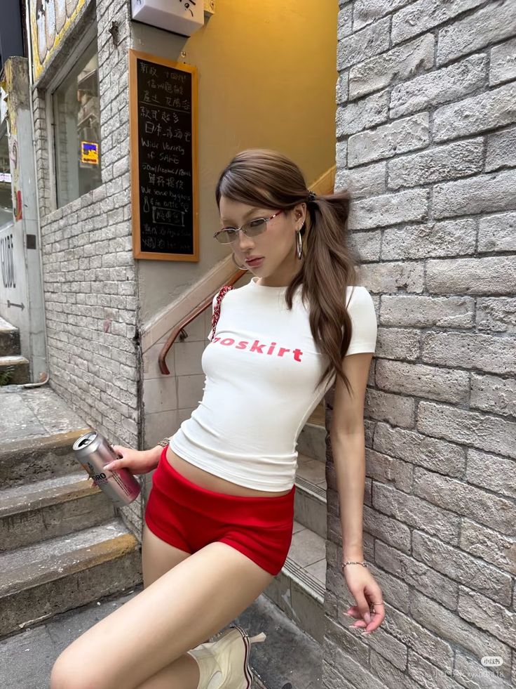 Woman in red shorts and a white t-shirt leaning against a brick wall