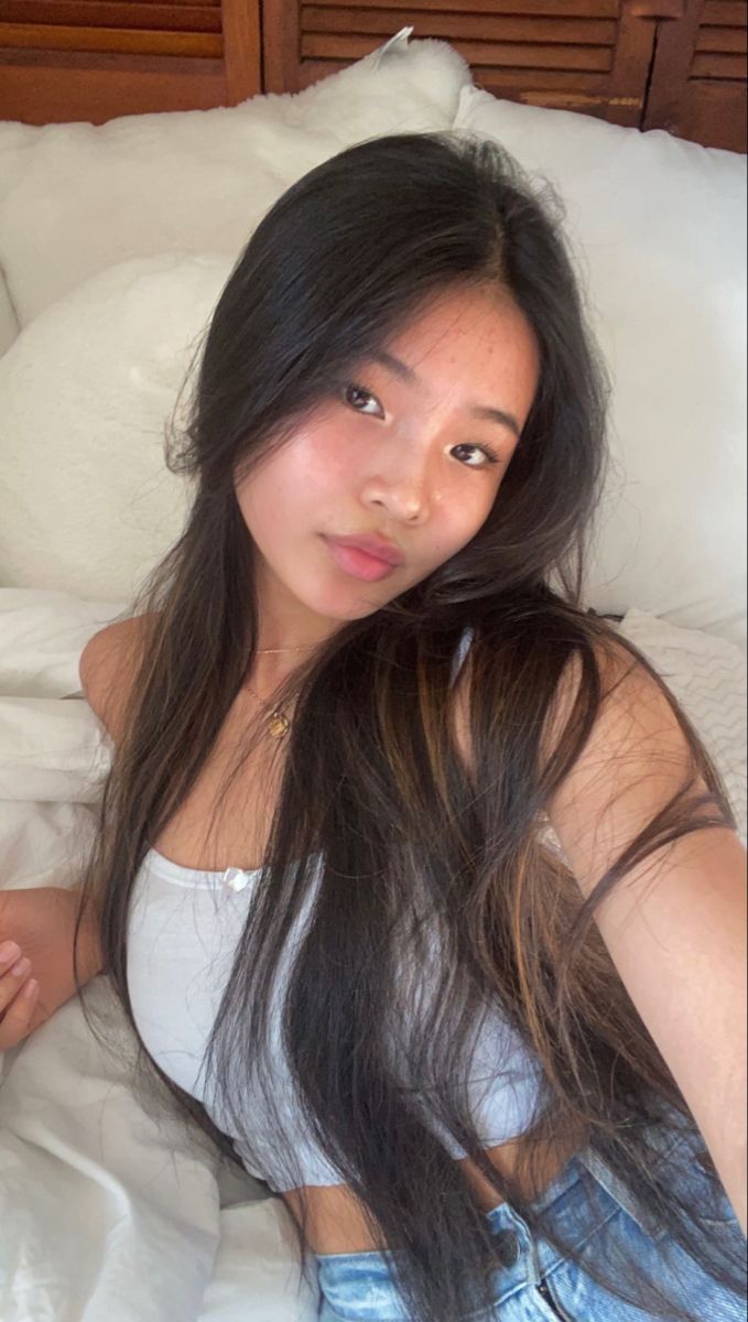 Woman with long dark hair wearing a white tank top and jeans, lying on a bed