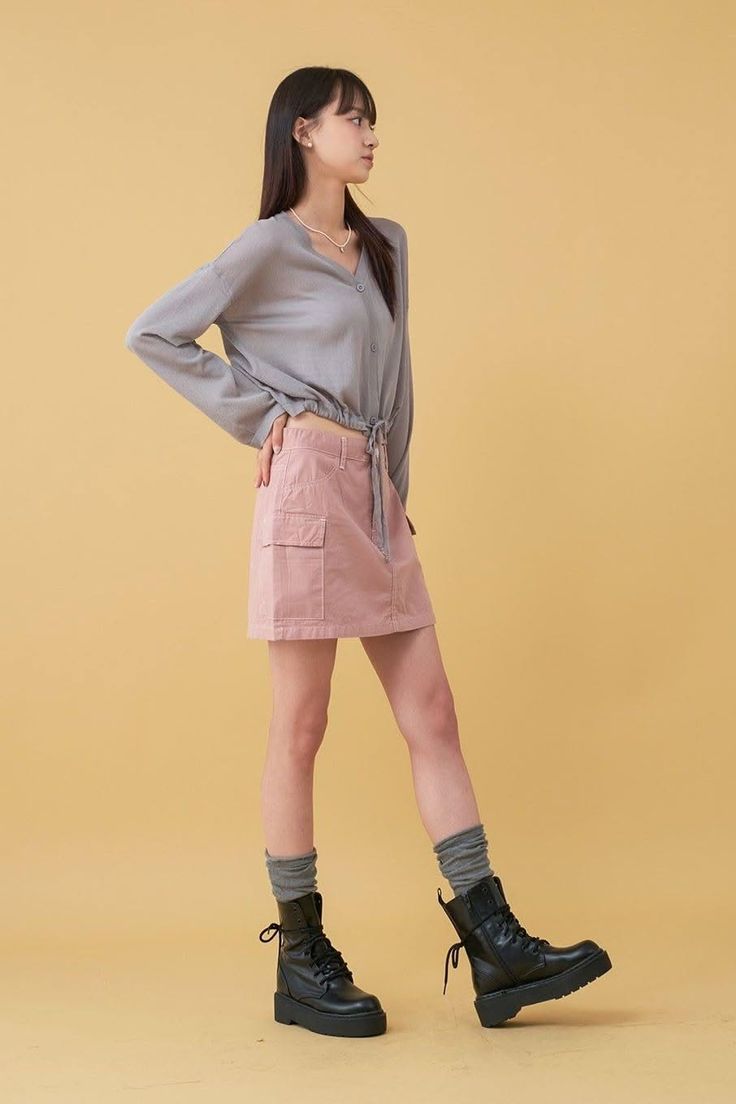 Woman in a gray shirt and pink skirt with combat boots posing against a yellow background