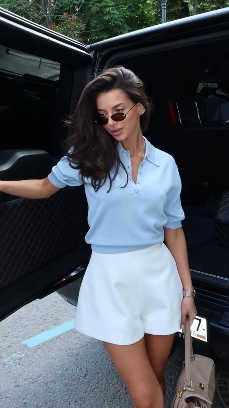 Woman with long hair and sunglasses wearing a polo shirt and white shorts next to a car