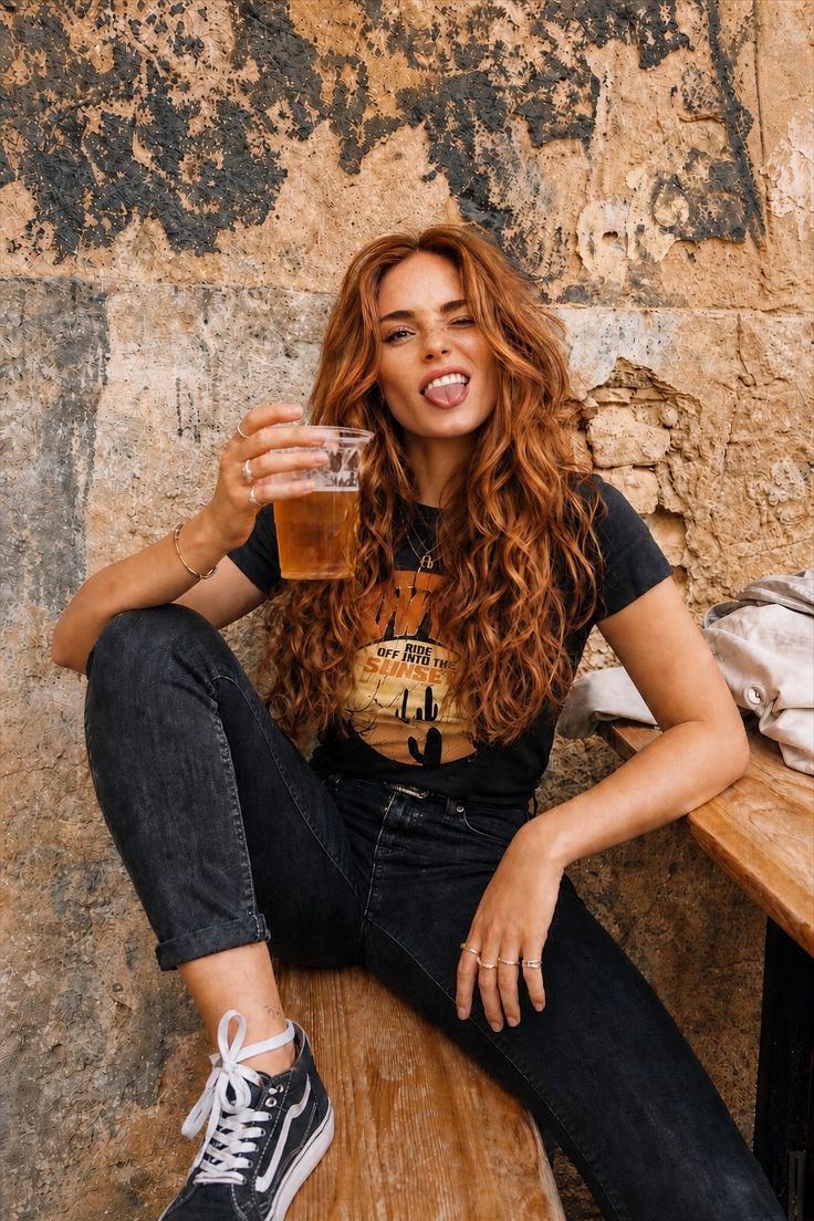 Woman with long curly hair holding a drink, sitting against a rustic wall
