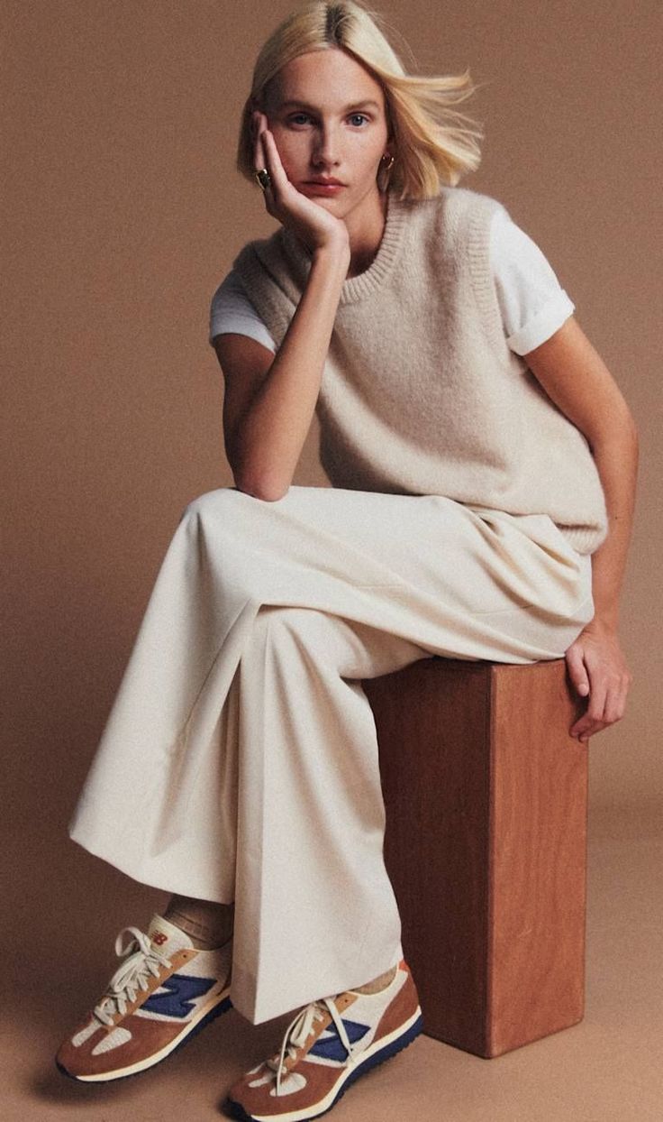 Woman with blonde hair wearing a sweater vest and wide-leg pants, sitting on a wooden block