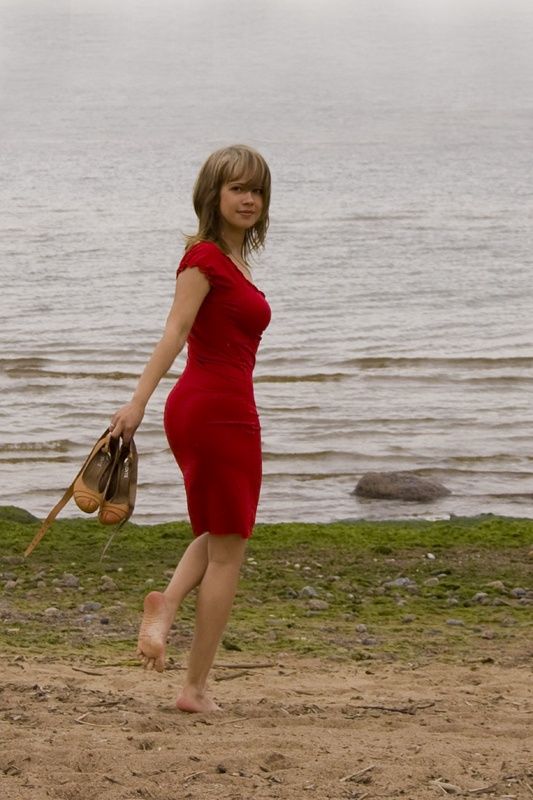 Woman in a red dress walking barefoot on a sandy beach, holding shoes