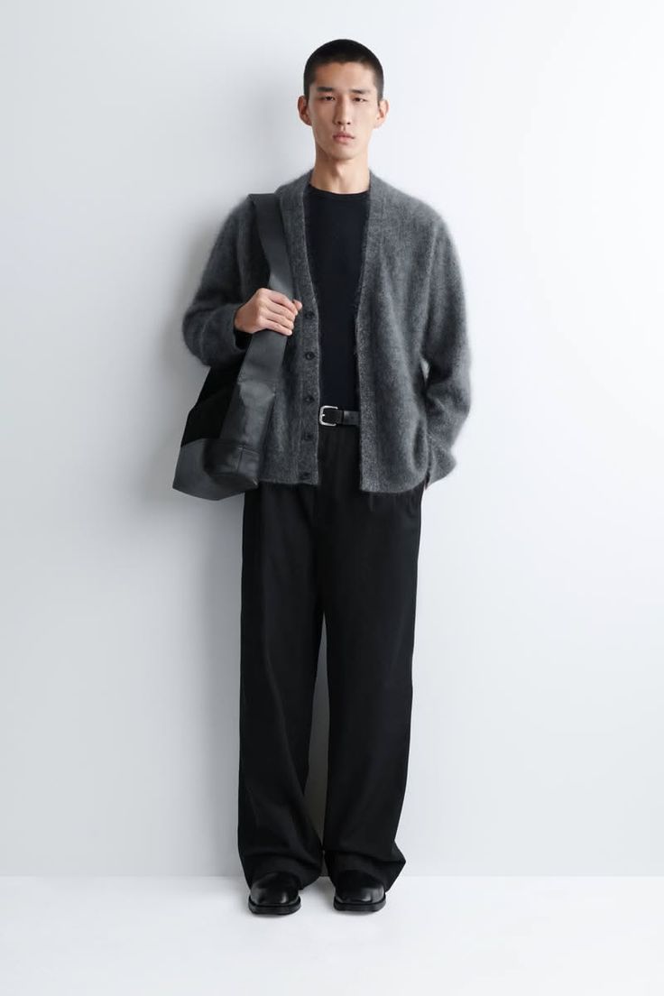 Man in a gray cardigan and black outfit posing against a white wall