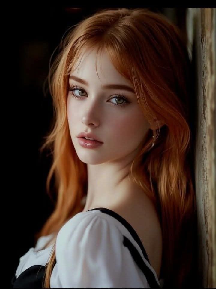 Woman with long red hair and green eyes in a white top, leaning against a wall
