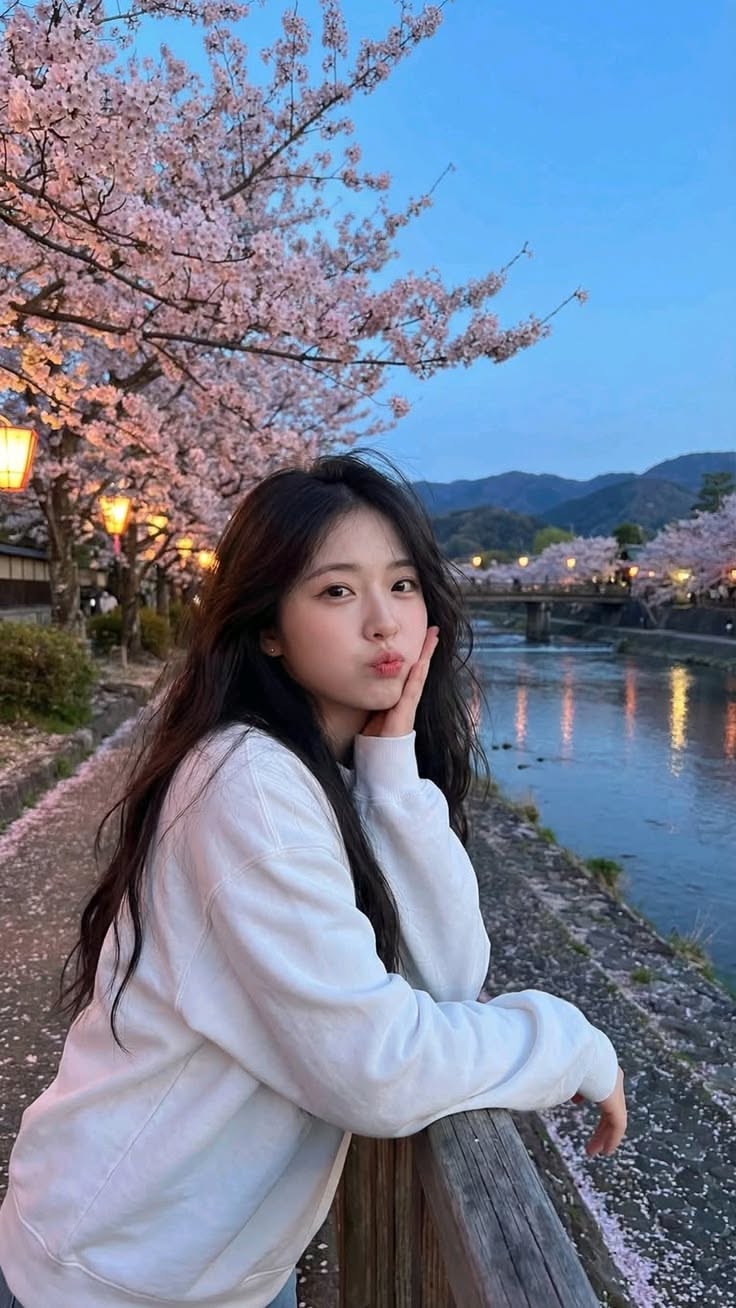 Woman in a white sweatshirt posing by a river with cherry blossoms at night