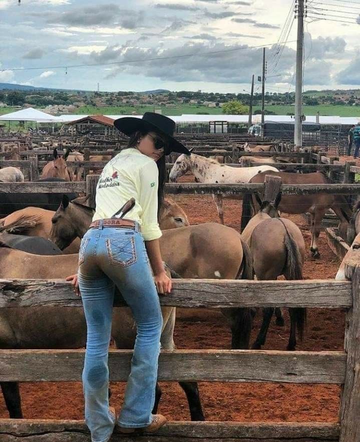 Woman in a cowgirl hat and muddy jeans leaning on a fence in a livestock pen