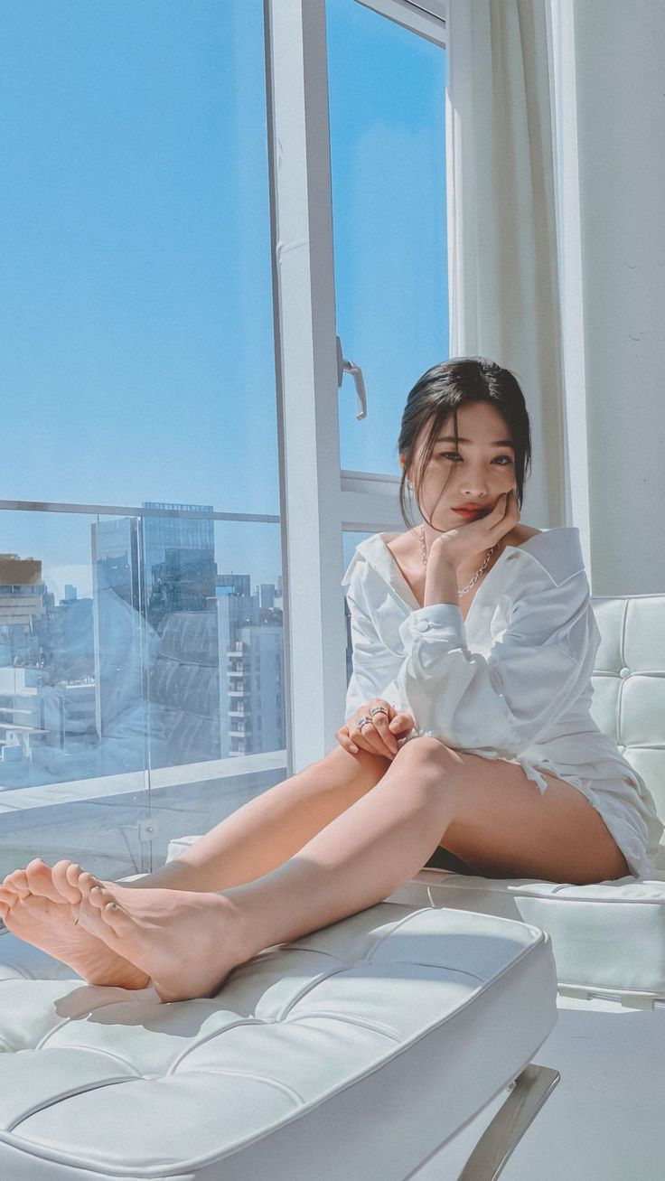 Woman in a white shirt dress sitting on a white couch by a window overlooking a cityscape