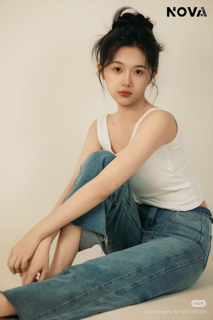 Young woman in a white tank top and blue jeans with a messy bun, sitting indoors