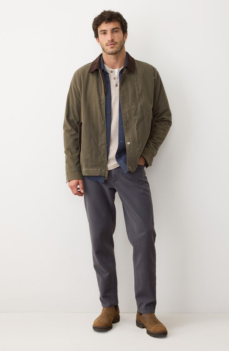 Man in an olive jacket, denim shirt, and dark pants standing against a white wall