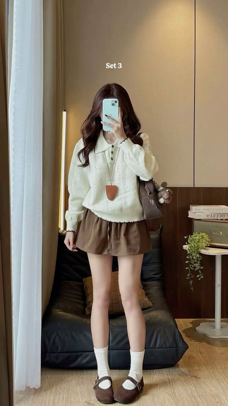 Woman in a white sweater and brown skirt taking a mirror selfie indoors