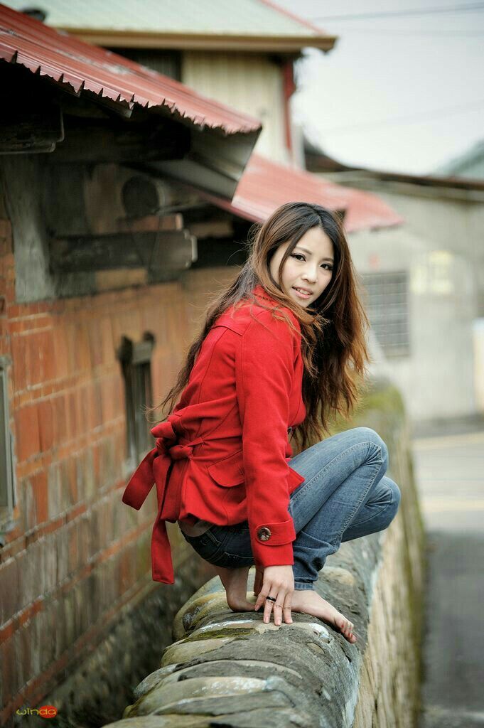 Woman in a red jacket and jeans squatting barefoot on a stone wall