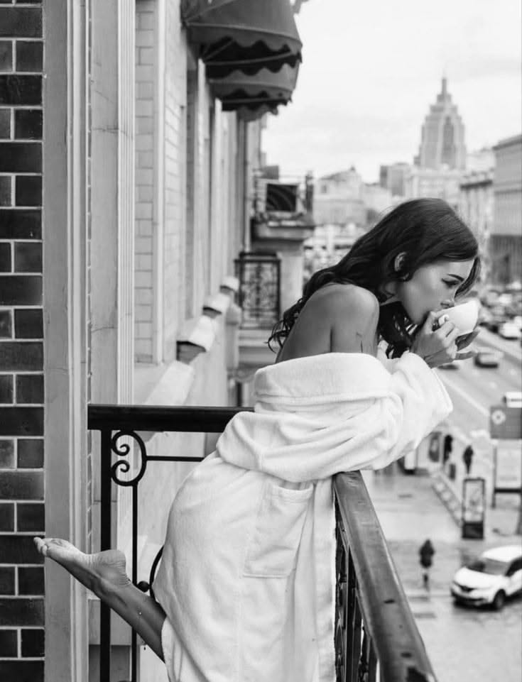 Woman in a white bathrobe drinking from a cup on a balcony overlooking a city