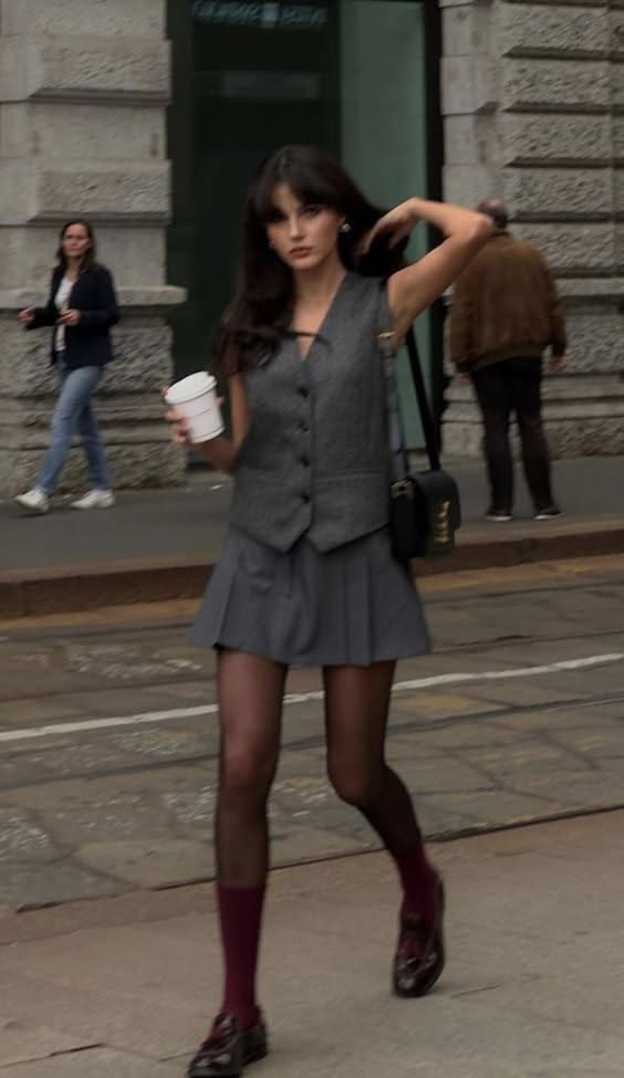 Woman in a grey vest and pleated skirt walking on a street with coffee