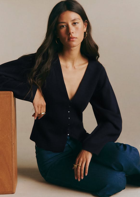 Woman with long dark hair wearing a deep v-neck top and jeans, sitting indoors