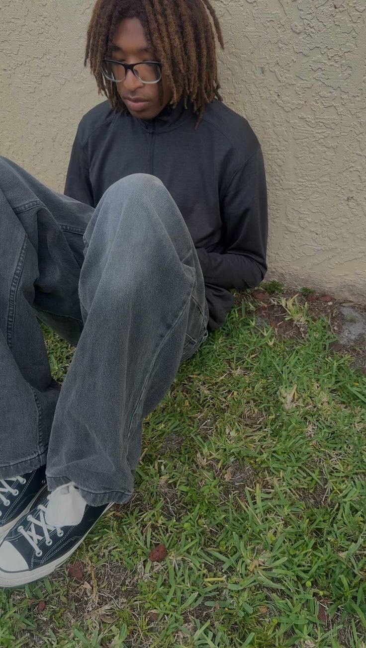 Person with dreadlocks and glasses sitting on grass next to a wall