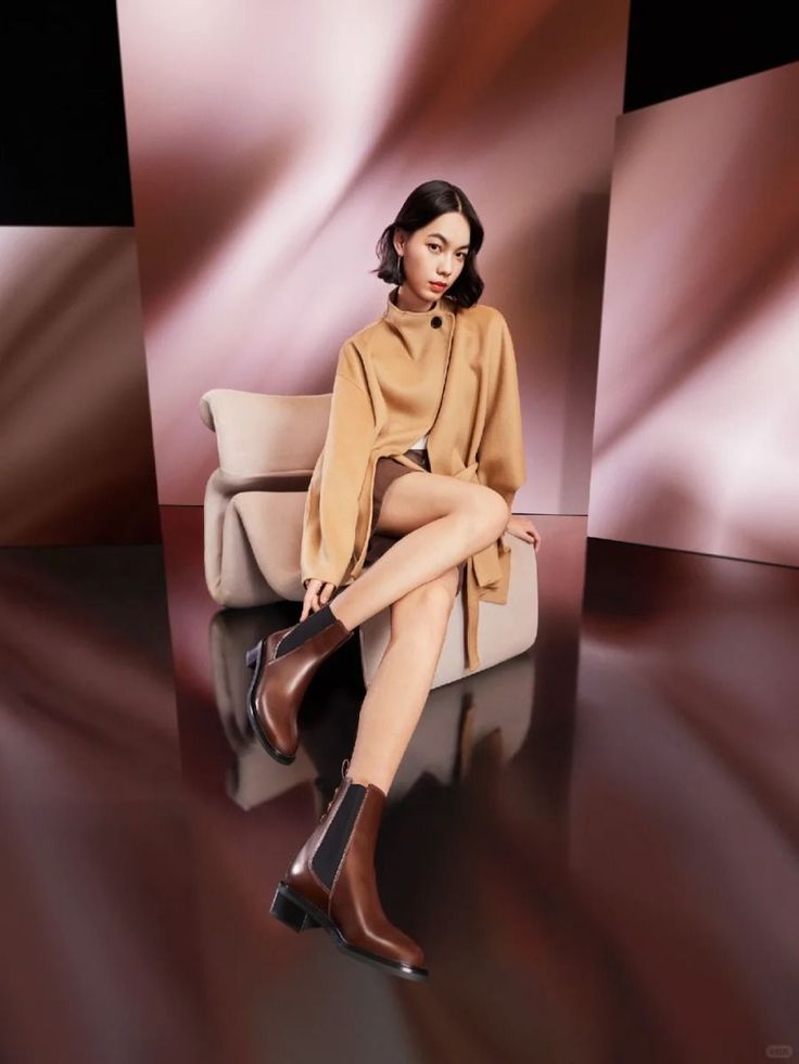 Woman with short hair wearing a camel coat and brown boots, sitting in a studio