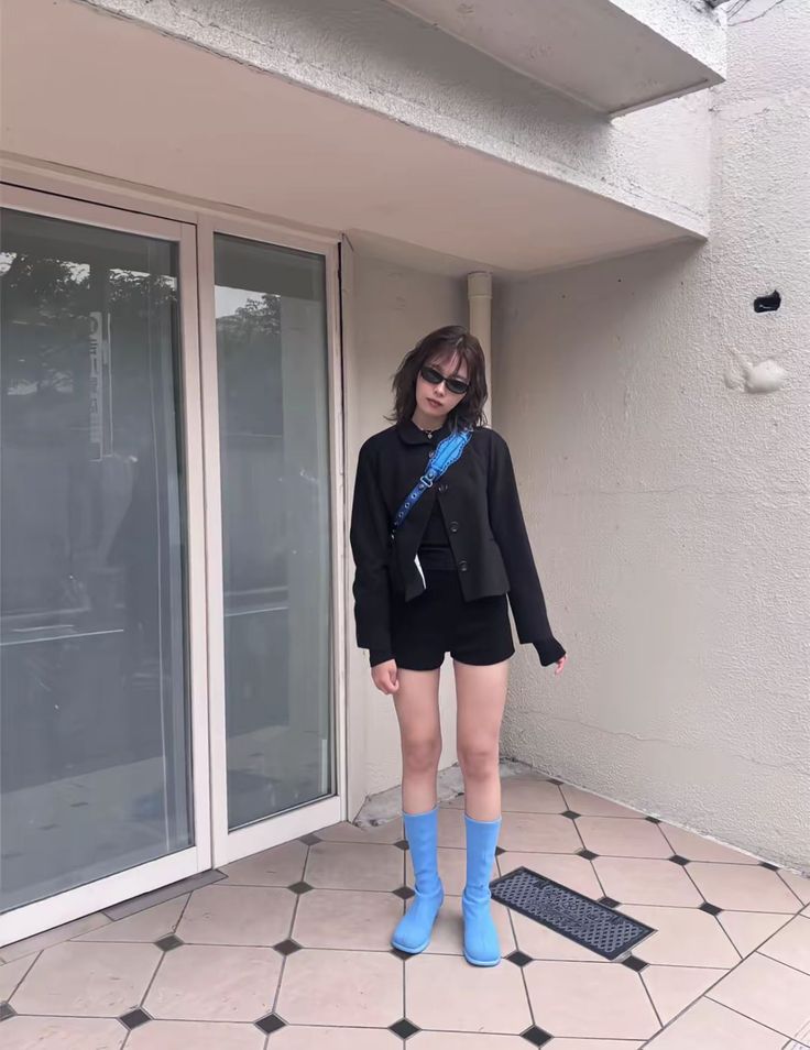 Woman in black jacket and shorts with blue boots and bag standing outdoors