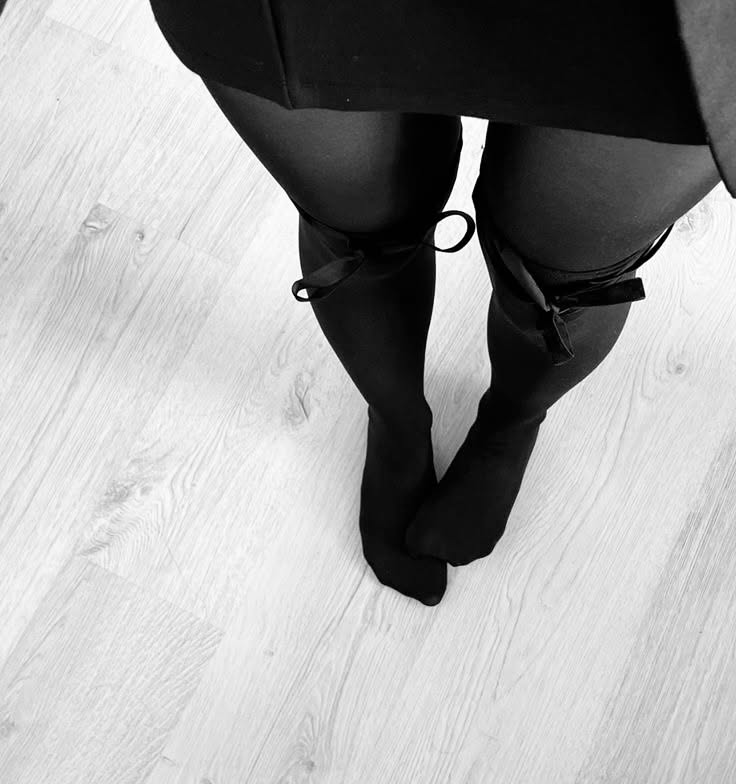 Person wearing black tights with bows and a black dress, standing on a wooden floor