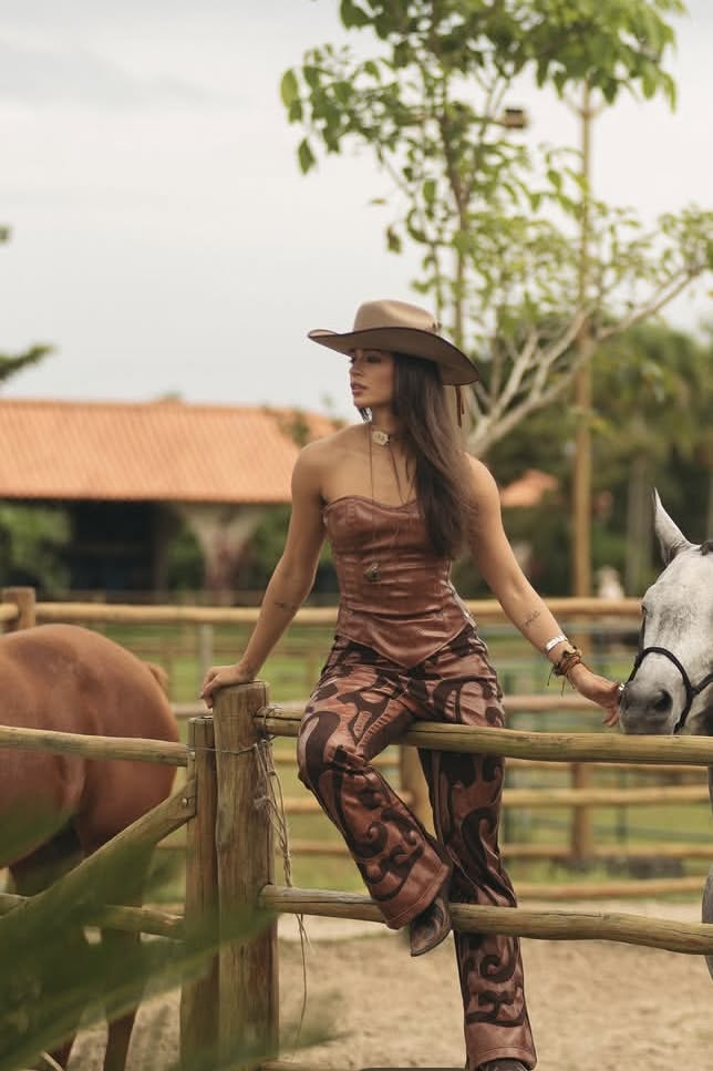 Woman in a cowboy hat and patterned outfit sitting on a fence at a ranch