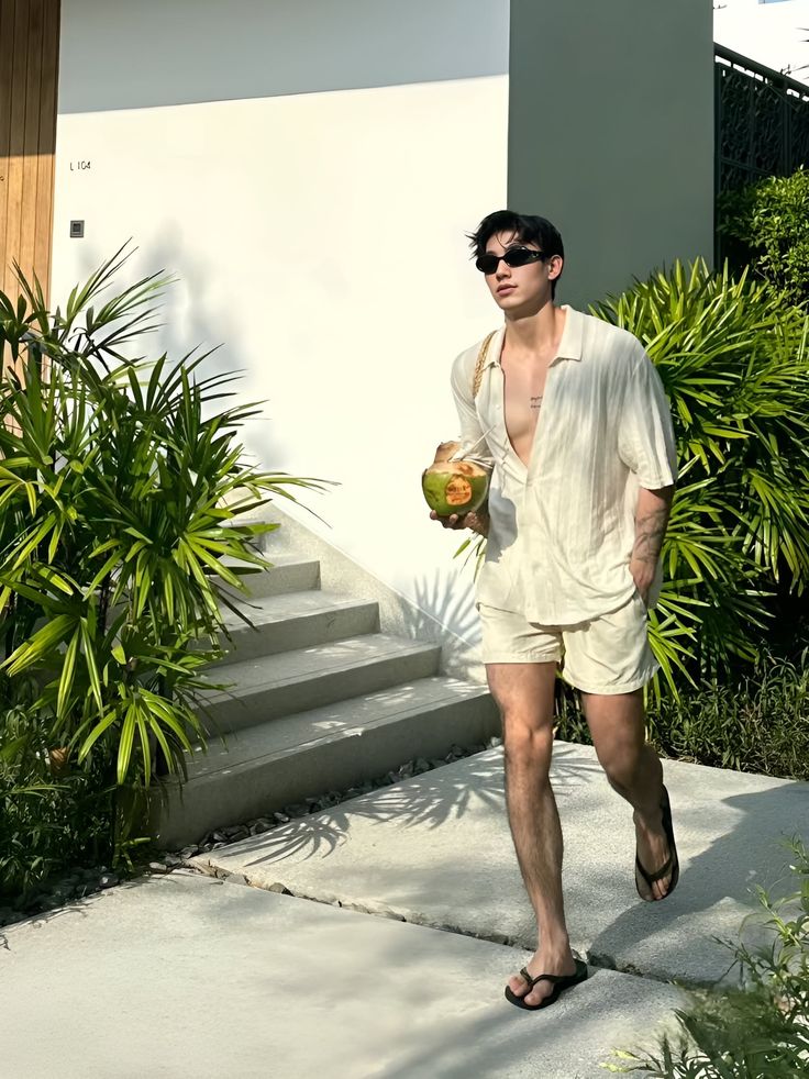 Man in an open shirt and shorts holding a coconut outdoors
