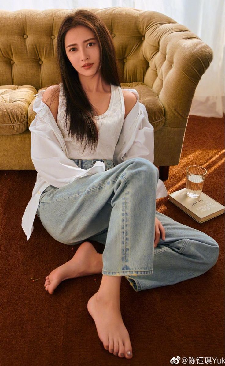 Woman with long hair wearing a white tank top and jeans, sitting on a brown carpet