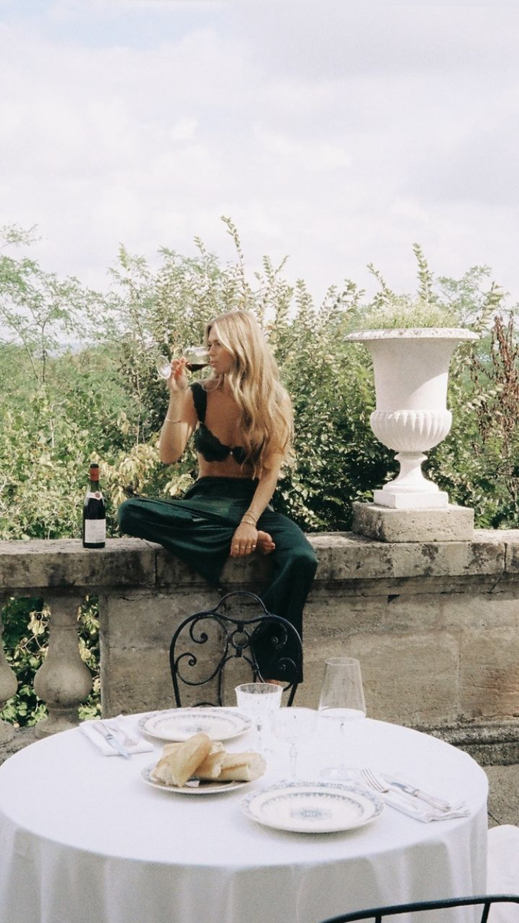 Woman in a green bralette and pants drinking wine on a stone wall