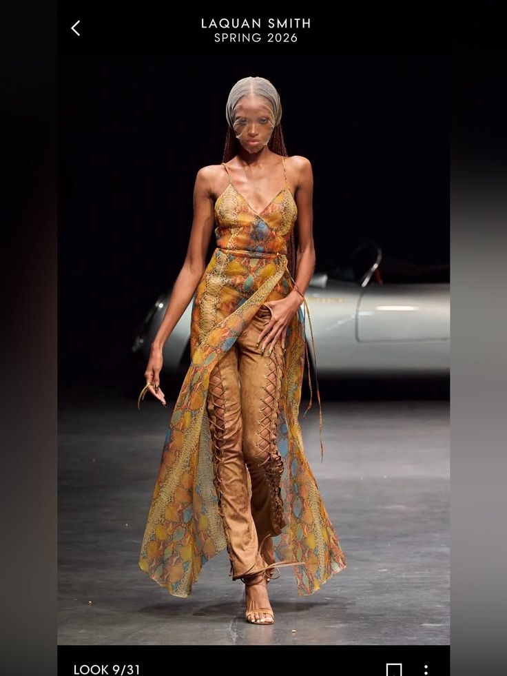 Model on a runway wearing a long sheer dress over lace-up pants