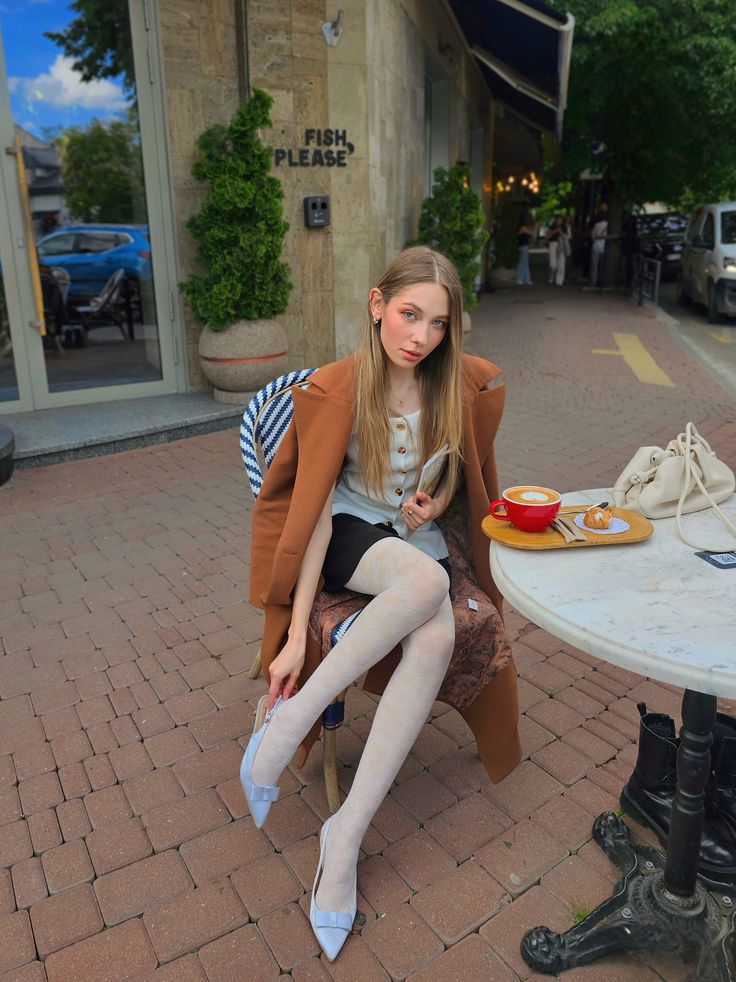 Woman in a brown coat and white tights sitting at an outdoor cafe table
