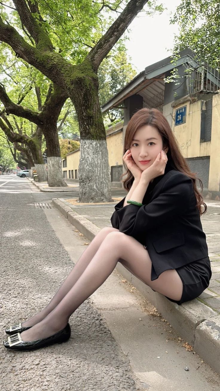 Woman in a black blazer and shorts sitting on a curb on a tree-lined street
