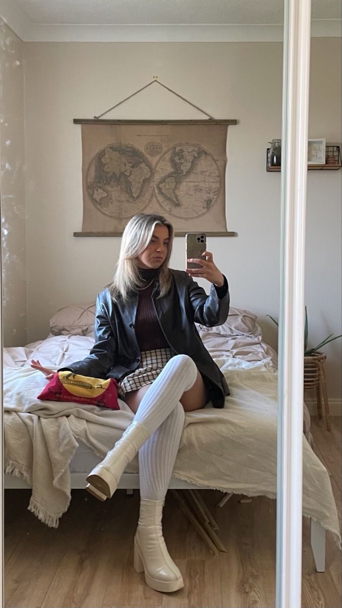 Woman in a leather jacket and plaid skirt taking a mirror selfie in a bedroom