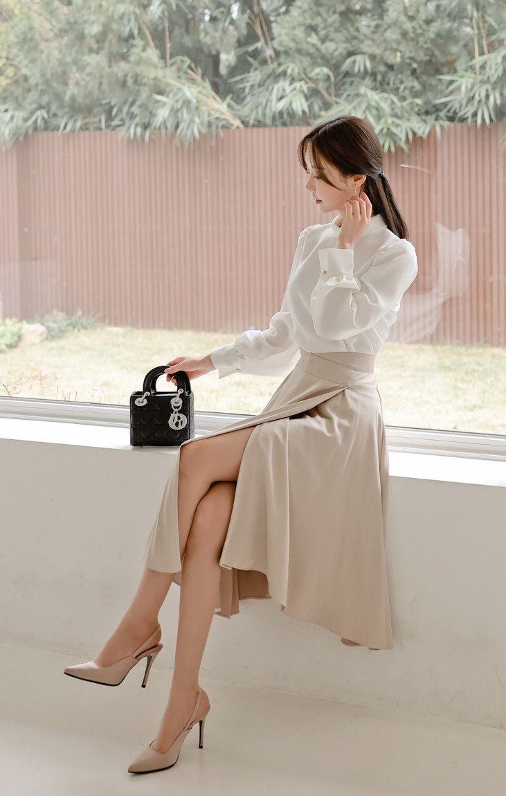 Woman in a white blouse and beige skirt sitting by a window with a handbag
