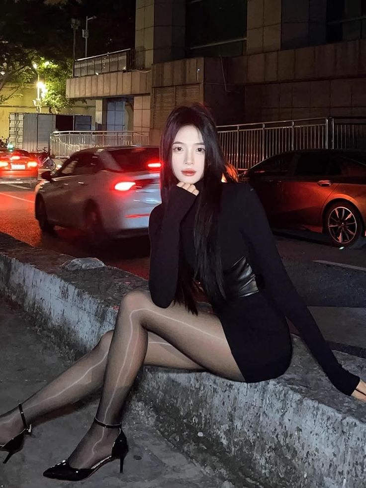 Woman in a black dress and sheer tights sitting on a curb at night