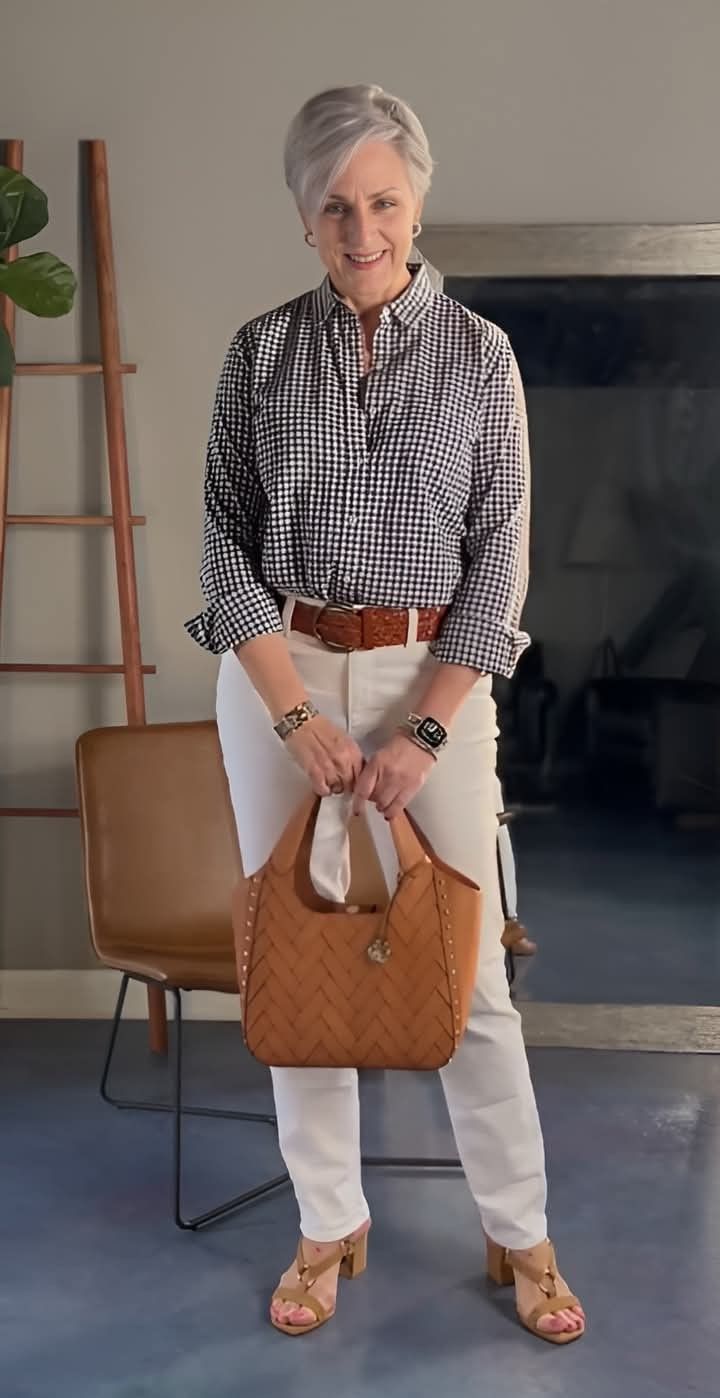Woman in a gingham shirt, white pants, and brown accessories standing indoors