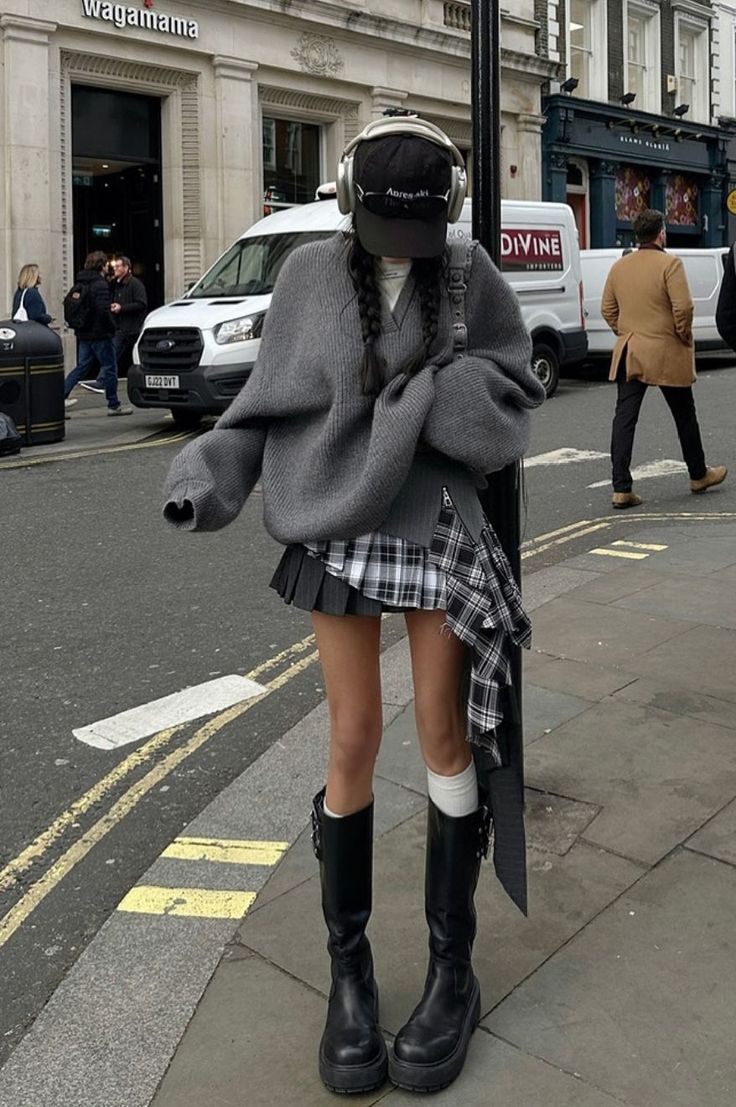 Person in a plaid skirt, oversized sweater, and knee-high boots on a city street