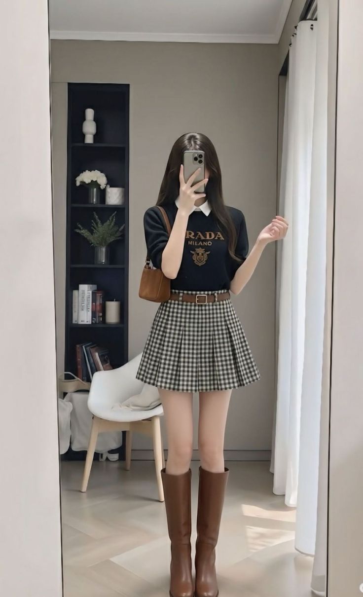Woman in a plaid skirt and brown boots taking a mirror selfie indoors