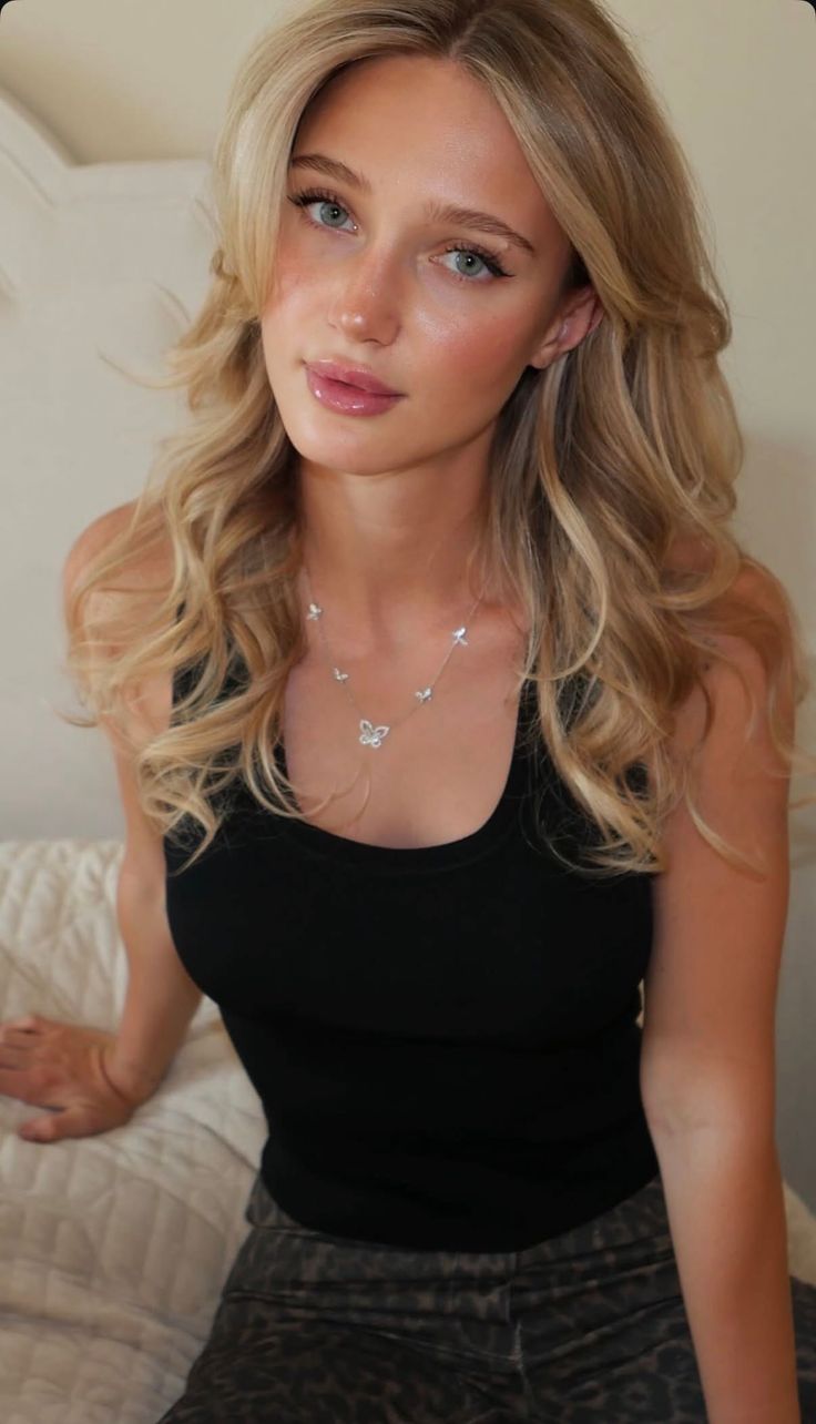 Blonde woman in a black tank top and butterfly necklace, looking at the camera