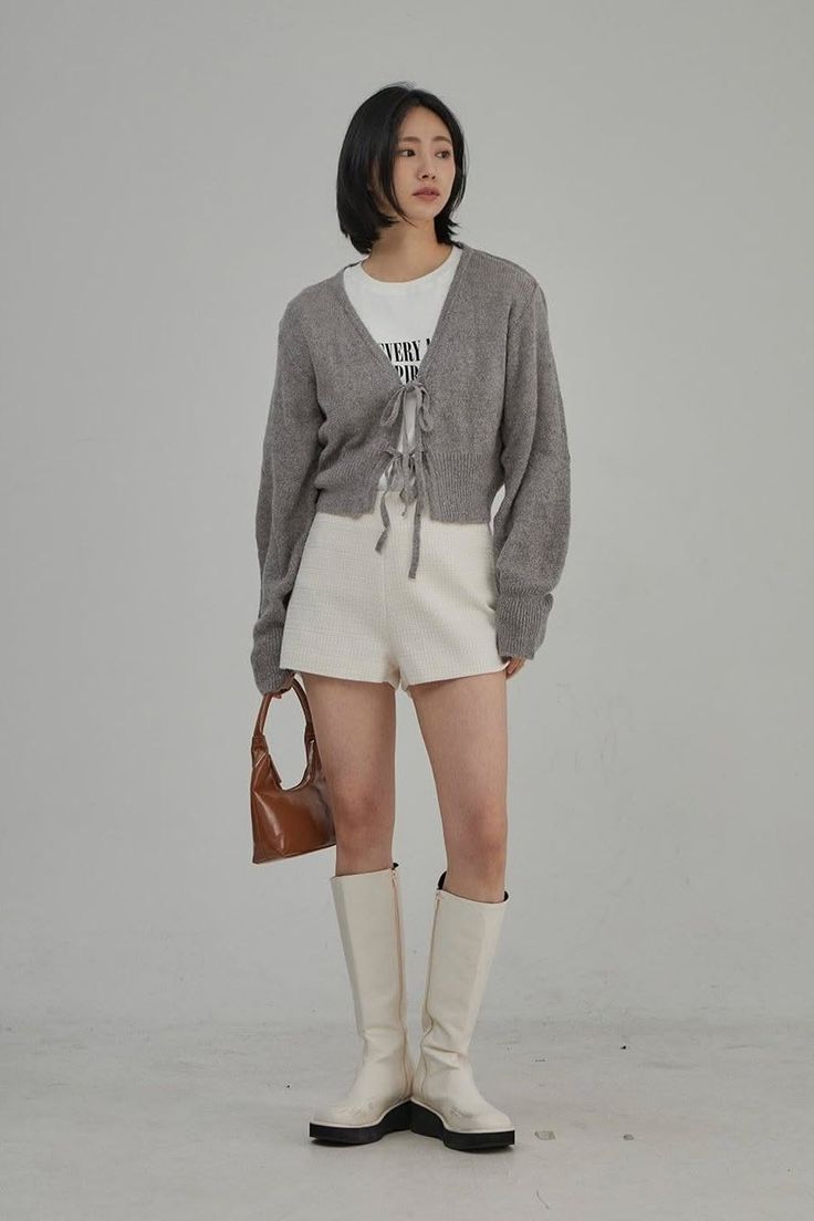 Woman in a gray cardigan, white shorts, and knee-high boots holding a brown handbag