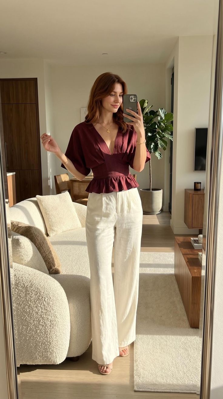 Woman in a maroon top and white pants taking a mirror selfie indoors