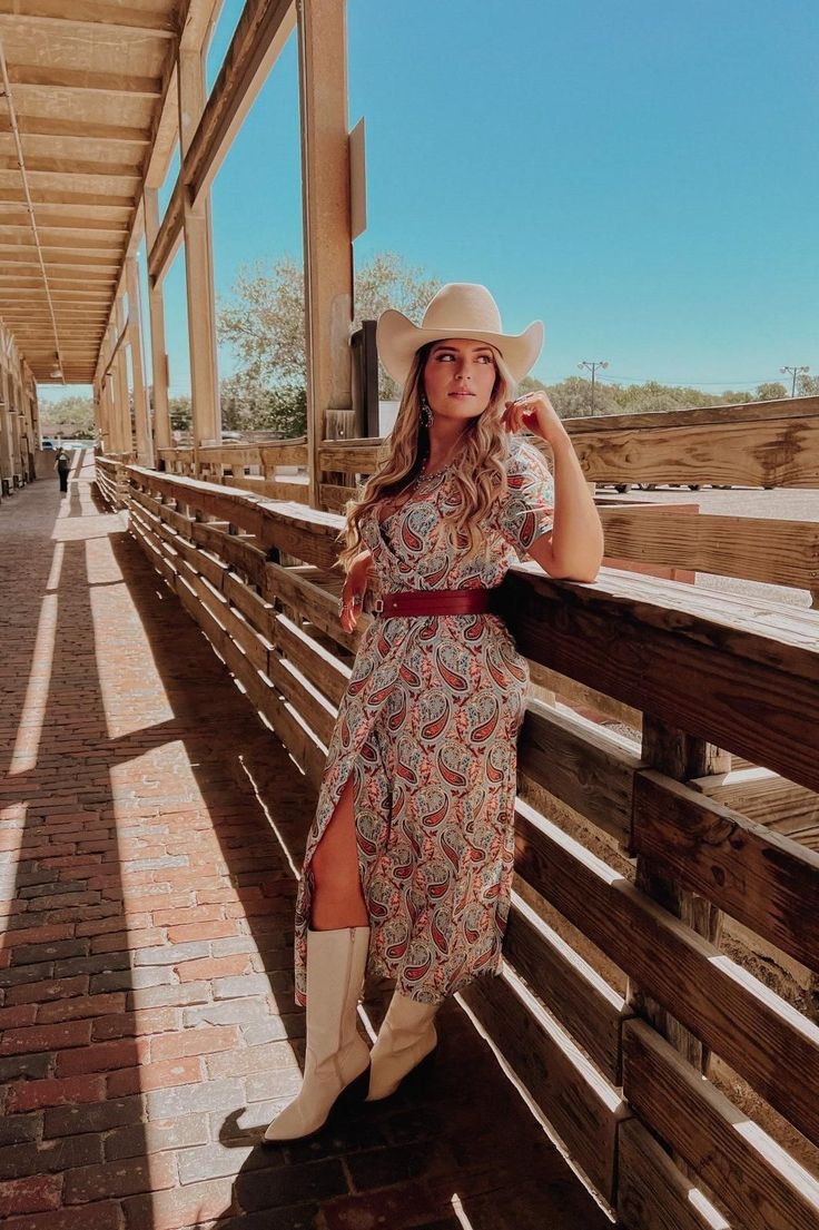 Woman in a paisley dress and cowboy hat leaning on a wooden fence outdoors