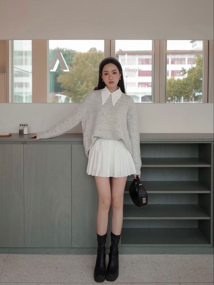 Woman in a grey sweater and white pleated skirt standing indoors
