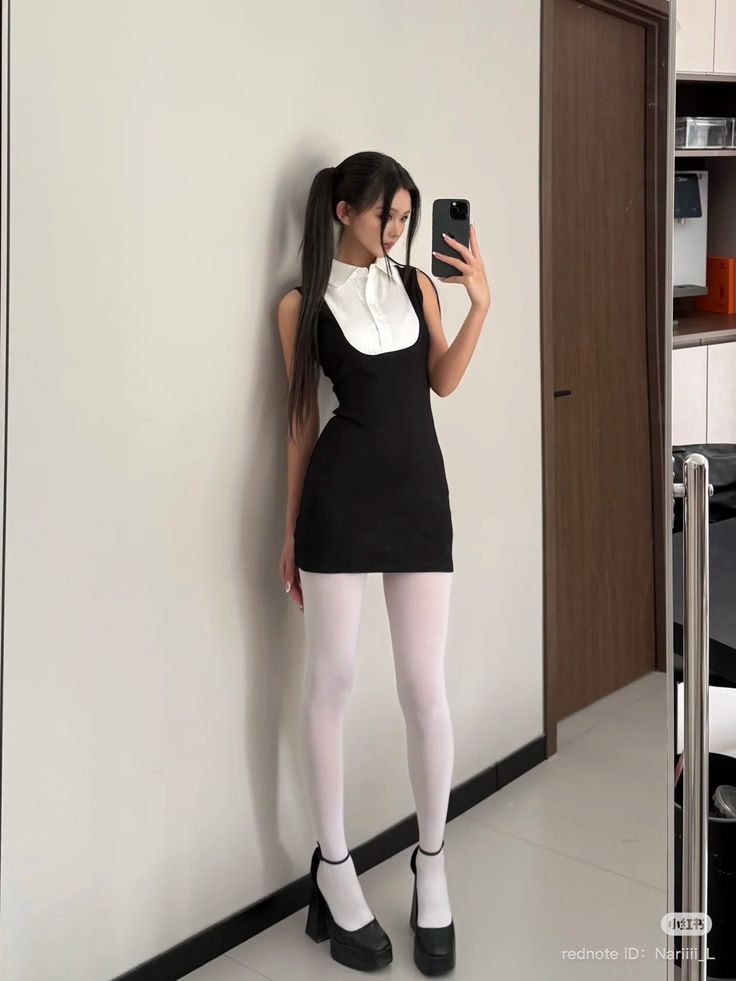 Woman in a black dress and white tights taking a mirror selfie