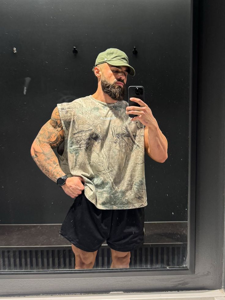 Muscular man with a beard and cap taking a mirror selfie in a camouflage shirt
