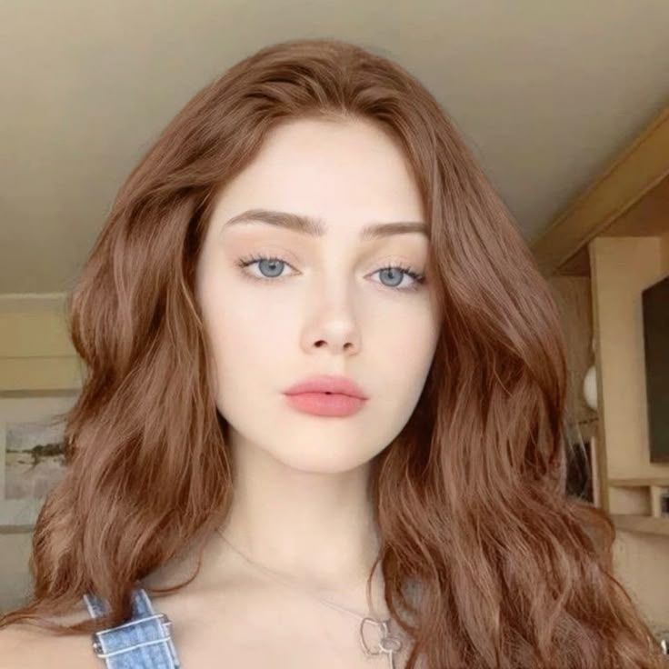 Woman with long red hair and blue eyes wearing denim overalls and a silver necklace