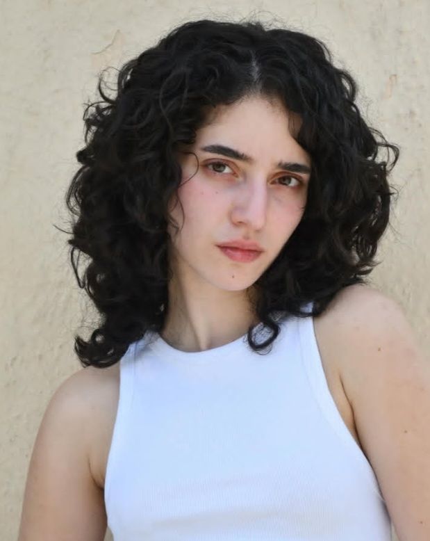 Woman with curly black hair wearing a white tank top against a plain background